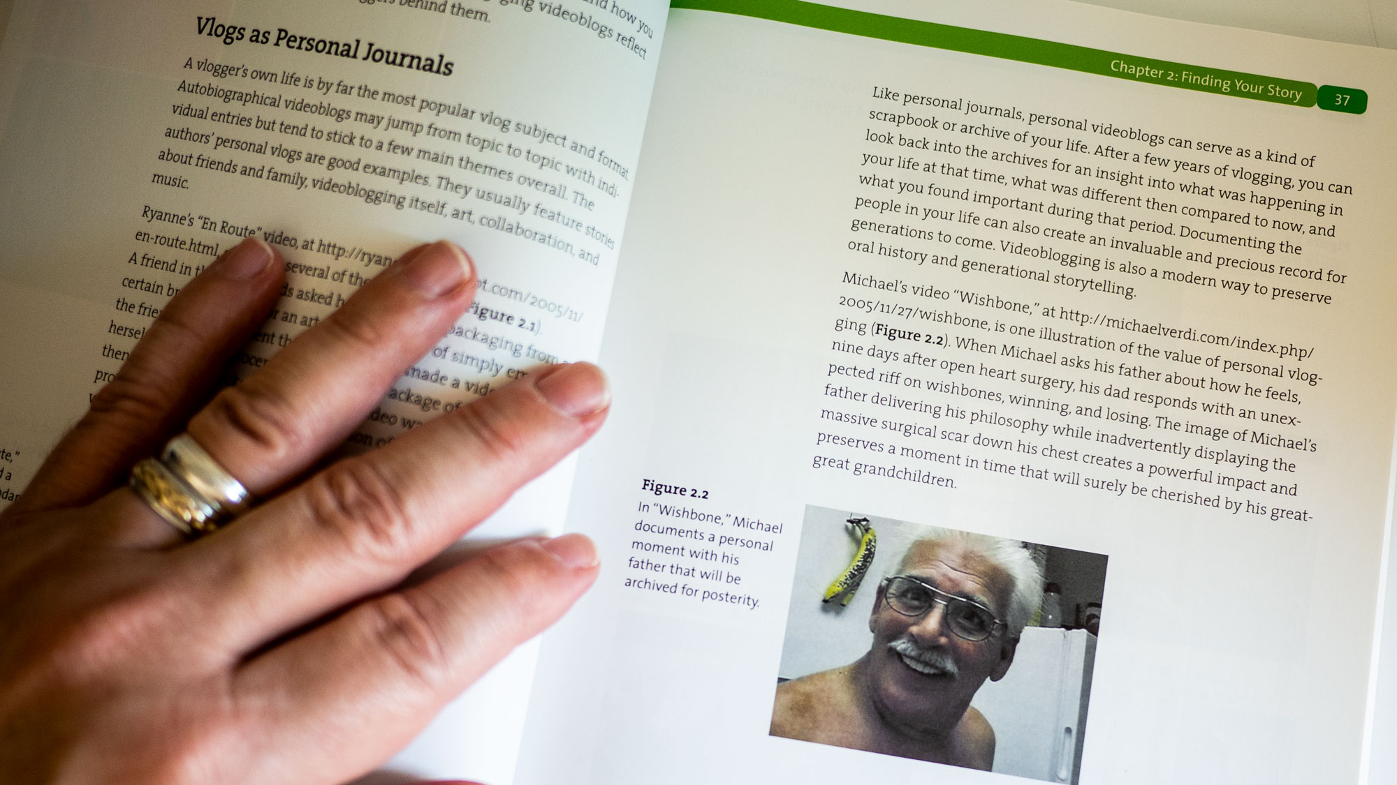 A hand holds open Secrets of Videoblogging to a page featuring a picture of a smiling man with gray hair and glasses. It's for a video entitled Wishbone.